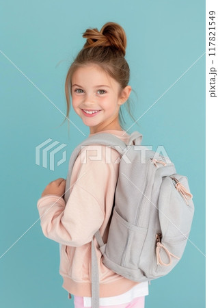 Smiling young girl with a grey backpack, wearing a light pink sweatshirt, posing against a blue background. Smiling young girl with a grey backpack, wearing a light pink sweatshirt, posing against a blue background. 117821549