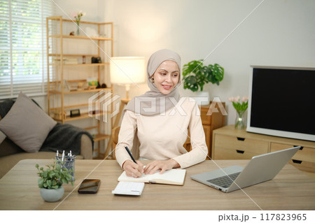 A woman wearing a head scarf is sitting at a desk with a laptop and a notebook 117823965