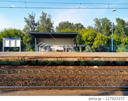 A platform for stopping electric trains for transporting passengers, an information board, rails, against the background of plants and the sky. City infrastructure, urbanism, transport, logistics A platform for stopping electric trains for transporting passengers, an information board, rails, against the background of plants and the sky. City infrastructure, urbanism, transport, logistics 117825257