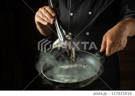 Cooking sprats fish in a frying pan. A chef fries capelin in a restaurant kitchen before dinner. Kitchen tongs with a small fish in the cook hand Cooking sprats fish in a frying pan. A chef fries capelin in a restaurant kitchen before dinner. Kitchen tongs with a small fish in the cook hand 117825818