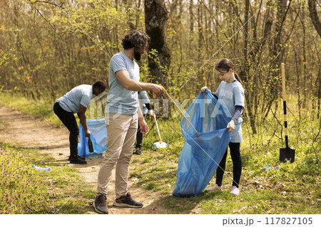 Team of activists gathering to clean up a forest natural area, using litter cleanup tools like garbage bags and a long claw to grab the junk. Volunteers collecting rubbish and recycling. Team of activists gathering to clean up a forest natural area, using litter cleanup tools like garbage bags and a long claw to grab the junk. Volunteers collecting rubbish and recycling. 117827105