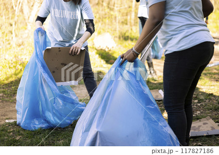 Women volunteers tidying the forest area of junk and plastic waste, collecting garbage with claw and blue disposal bag. Team of activists volunteering to preserve natural environment. Women volunteers tidying the forest area of junk and plastic waste, collecting garbage with claw and blue disposal bag. Team of activists volunteering to preserve natural environment. 117827186