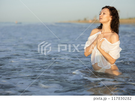 Topless woman in white shirt kneeling on coast 117830282