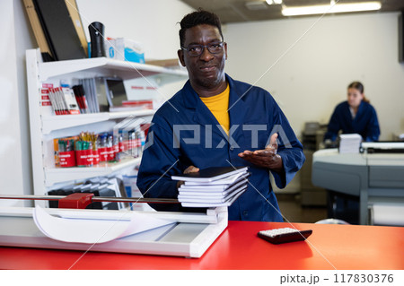 Portrait of friendly male employee of printing house with stack of notebooks in her hands Portrait of friendly male employee of printing house with stack of notebooks in her hands 117830376