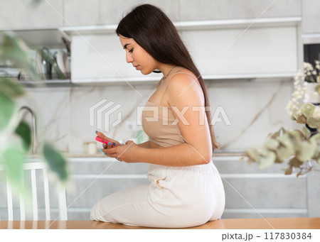 Young woman sitting on kitchen table and looking at her phone Young woman sitting on kitchen table and looking at her phone 117830384