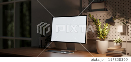 A close-up image of a computer with a white screen mockup on a wooden table in a dark room at night. 117830399