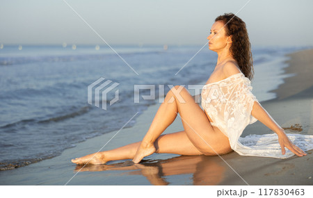 Adult woman in white peignoir sitting on coast Adult woman in white peignoir sitting on coast 117830463
