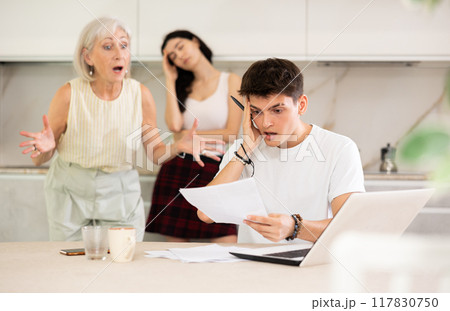 Young guy while working on laptop listens to claims of elderly mother and young sister girl Young guy while working on laptop listens to claims of elderly mother and young sister girl 117830750