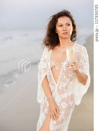 Portrait of woman in white mesh cape with glass of champagne posing on seashore 117830785