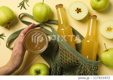 Bottles and glass with cider, hand, green apples and bag on beige background, top view Bottles and glass with cider, hand, green apples and bag on beige background, top view 117833972