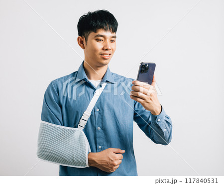 Injured man smiles with a broken arm in a splint, holding a smartphone. Happy Asian woman with a supported hand in a sling on white background. Social media and space for text. Injured man smiles with a broken arm in a splint, holding a smartphone. Happy Asian woman with a supported hand in a sling on white background. Social media and space for text. 117840531