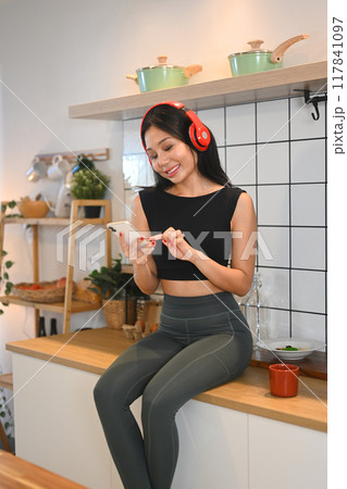 Happy young fitness woman listening music through headphones and using mobile phone in kitchen 117841097