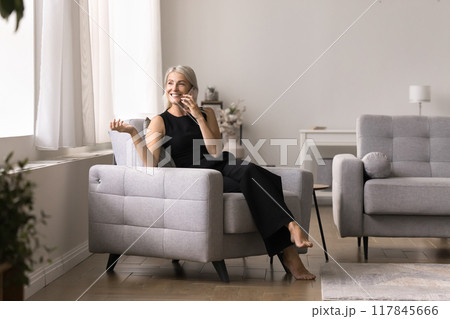 Happy mature woman talking on mobile phone sitting on armchair Happy mature woman talking on mobile phone sitting on armchair 117845666
