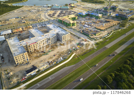 View from above of unfinished frame of apartment condos with wooden roof beams under construction. Development of residential housing near state highway in US suburbs. Real estate market in the USA View from above of unfinished frame of apartment condos with wooden roof beams under construction. Development of residential housing near state highway in US suburbs. Real estate market in the USA 117846268
