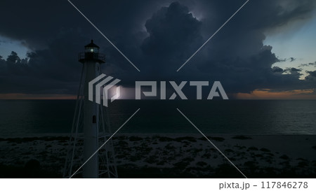 White tall lighthouse on sea shore with blinking light at stormy night for commercial vessels navigation. Thunderstorm with lightnings over ocean water posing danger for ships White tall lighthouse on sea shore with blinking light at stormy night for commercial vessels navigation. Thunderstorm with lightnings over ocean water posing danger for ships 117846278