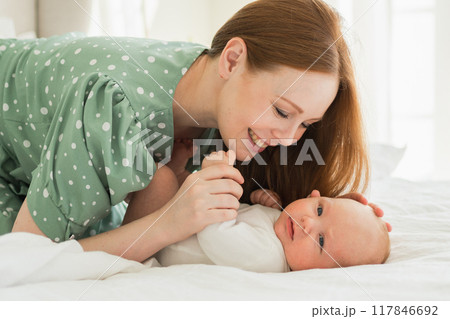 Happy family at home. Mother playing with little toddler child on bed. Mom baby relax playing having fun together. Mother looks at baby with love care. Mom of breast feeding baby rest in living room Happy family at home. Mother playing with little toddler child on bed. Mom baby relax playing having fun together. Mother looks at baby with love care. Mom of breast feeding baby rest in living room 117846692