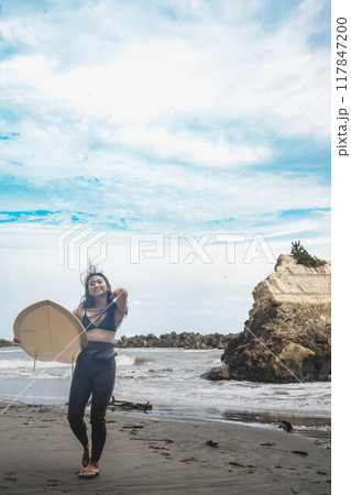 風が吹く海でサーフボードを持って立っている若い女性 117847200