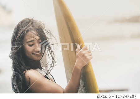 風が吹く海でサーフボードを持って立っている若い女性 117847207
