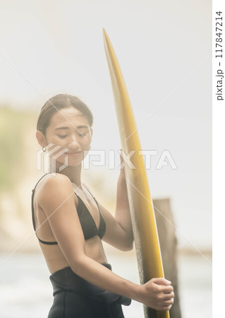 風が吹く海でサーフボードを持って立っている若い女性 風が吹く海でサーフボードを持って立っている若い女性 117847214