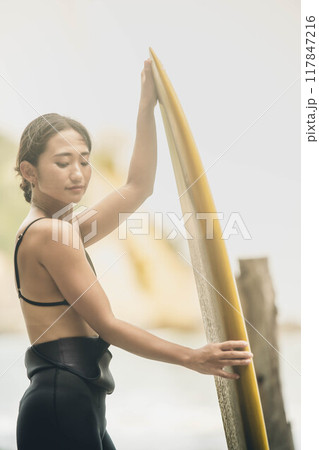 風が吹く海でサーフボードを持って立っている若い女性 風が吹く海でサーフボードを持って立っている若い女性 117847216