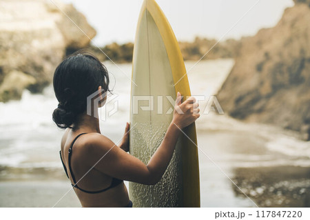 風が吹く海でサーフボードを持って立っている若い女性 117847220
