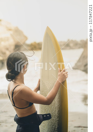 風が吹く海でサーフボードを持って立っている若い女性 117847221