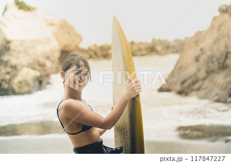 風が吹く海でサーフボードを持って立っている若い女性 117847227