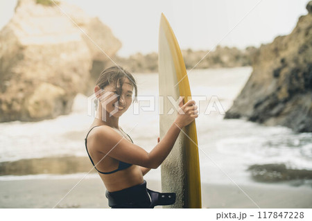 風が吹く海でサーフボードを持って立っている若い女性 117847228