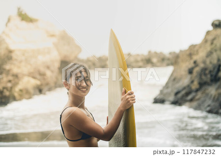 風が吹く海でサーフボードを持って立っている若い女性 風が吹く海でサーフボードを持って立っている若い女性 117847232