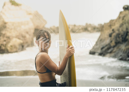 風が吹く海でサーフボードを持って立っている若い女性 117847235