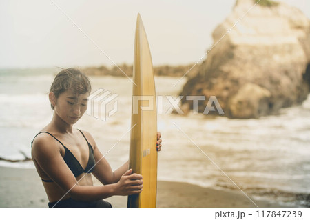 風が吹く海でサーフボードを持って立っている若い女性 風が吹く海でサーフボードを持って立っている若い女性 117847239