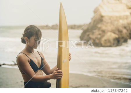風が吹く海でサーフボードを持って立っている若い女性 風が吹く海でサーフボードを持って立っている若い女性 117847242