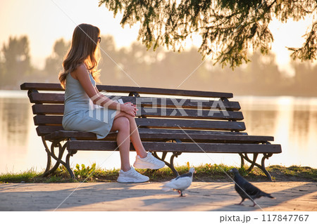 Lonely woman sitting on lake side bench enjoying warm summer evening. Solitude and relaxation concept. 117847767