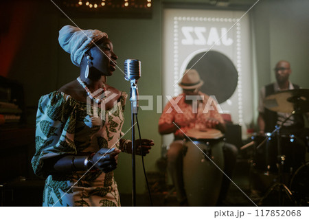 Young Black woman wearing stylish dress with headwrap singing in microphone during performance with jazz band 117852068