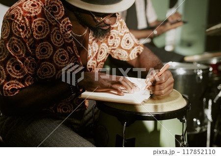 African American musician sitting at tumbadora writing note in notebook during rehearsal with jazz band African American musician sitting at tumbadora writing note in notebook during rehearsal with jazz band 117852110
