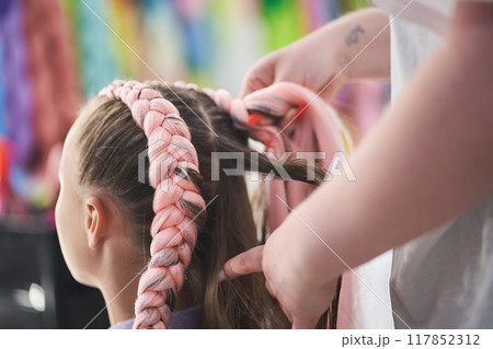 Close-up, side view. The hands of the master weave the kanekalon material into the hair. The master braids the child's pigtails from a pink kanekalon. High quality photo Close-up, side view. The hands of the master weave the kanekalon material into the hair. The master braids the child's pigtails from a pink kanekalon. High quality photo 117852312
