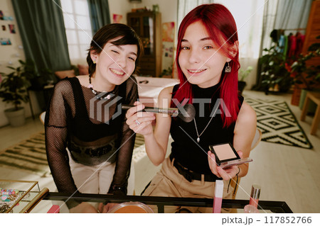 Two friends happily sharing makeup tips while sitting in cozy and well-decorated living room surrounded by mirrors, brushes, and cosmetics, creating a joyful and friendly atmosphere 117852766