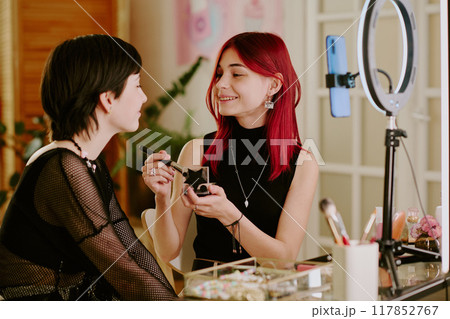 Two young women engaging in beauty session indoors, one applying makeup to other while sitting in modern studio setting with professional equipment visible around them 117852767