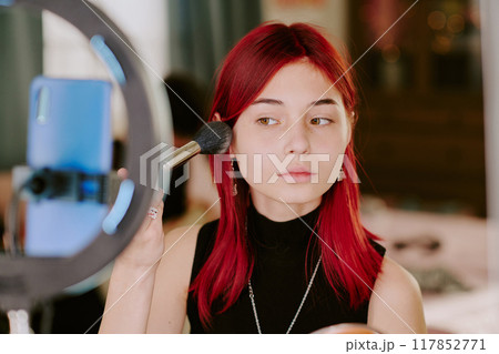 Young woman with red hair applying makeup with brush using cosmetic mirror with ring light for precise application and even blending 117852771