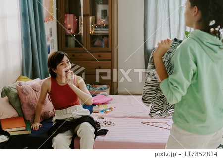 Young woman sitting on bed, wearing red tank top, talking to friend who is holding a patterned shirt in cozy bedroom full of books and personal items Young woman sitting on bed, wearing red tank top, talking to friend who is holding a patterned shirt in cozy bedroom full of books and personal items 117852814