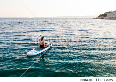A woman paddle boarding on tranquil waters near a rocky shoreline at sunset. 117855069