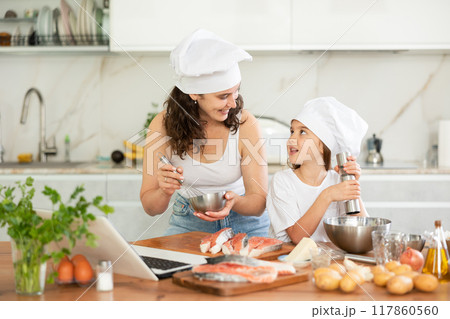 Happy mother and daughter wearing chef hats preparing salmon in the kitchen Happy mother and daughter wearing chef hats preparing salmon in the kitchen 117860560