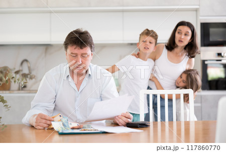 Sad father looks at bills and counts money. Upset wife with children in background 117860770