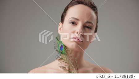 Beauty, health, cosmetics, anti-aging therapy and skin care concept - young beautiful brunette Caucasian woman holding a colorful peacock feather, concept of tenderness, softness and lightness 117861887