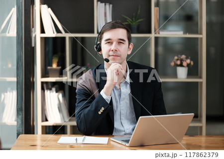 A man in a suit is sitting at a desk with a laptop and a headset 117862379