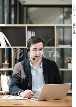 A man wearing a suit and a headset is sitting at a desk with a laptop 117862380