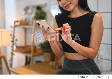 Smiling young fitness woman listening music through headphones and using mobile phone in kitchen Smiling young fitness woman listening music through headphones and using mobile phone in kitchen 117863294