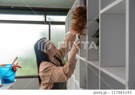 Diligent housekeeper cleaning high office shelves with feather duster Diligent housekeeper cleaning high office shelves with feather duster 117865243