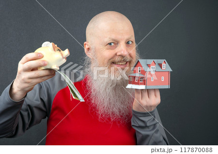 bald man with a long gray beard holding a piggy bank with a dollar bill and a small model house bald man with a long gray beard holding a piggy bank with a dollar bill and a small model house 117870088