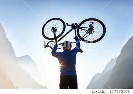 male cyclist with a raised bicycle against the backdrop of the sun and mountains male cyclist with a raised bicycle against the backdrop of the sun and mountains 117870198